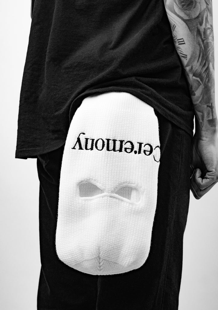 Ceremony White Balaclava hanging upside down from the pant pocket on person from side view featuring black embroidered Ceremony Logo